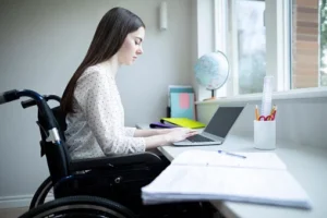 A young woman in a wheelchair working on a laptop at a desk by a window. She is wearing a white blouse and focused on the screen, with stationery items visible on the desk.