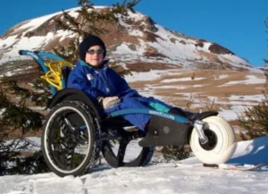A child wearing a blue jacket and sunglasses sits in a specialized wheelchair on snowy ground, with a mountainous landscape in the background. The wheelchair is designed for outdoor use.