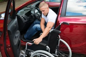 A man in a white shirt is placing a wheelchair into the back seat of a red car, while seated inside the vehicle. The scene takes place outdoors with sunlight visible.