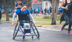 A woman in a wheelchair participating in a race, concentrating on the path ahead. Several runners are visible in the background, with trees lining the side of the road.