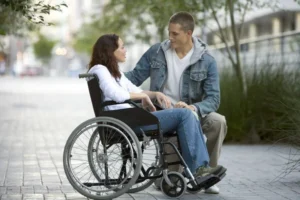 A young woman in a wheelchair conversing with a young man seated beside her on a pathway lined with greenery. Both are wearing casual clothing, and the scene is set in an outdoor urban environment.
