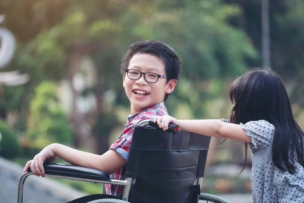 A smiling boy in a wheelchair enjoys time outdoors while a girl pushes him from behind in a playful manner. Lush greenery and park features are visible in the background.