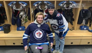 Two young men in a hockey locker room. One man, in a wheelchair, wears a Winnipeg Jets jersey, while the other, standing beside him, wears a similar jersey and a hat. Hockey equipment is visible in the background.