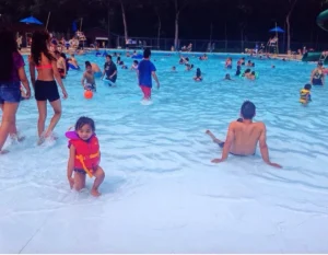 A vibrant outdoor pool scene at Kildonan Park, featuring families and children swimming and playing in the water. A child in a life jacket is smiling and playing near the edge, while adults relax and socialize in the background. The setting includes a gradual slope entry for accessibility.