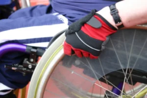 A close-up of a gloved hand gripping the wheel of a wheelchair, with a focus on the wheel's rim and the textured grip of the glove. The wheelchair has a purple frame and is set against a blurred background.