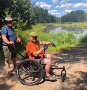 A person sitting in an all-terrain wheelchair is being pushed along a trail by another person. The scene features a lush green landscape with trees and a pond in the background under a sunny sky.