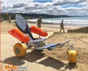 A beach wheelchair with flotation devices, designed for accessibility, parked on sandy terrain near the ocean. In the background, people are walking along the beach.