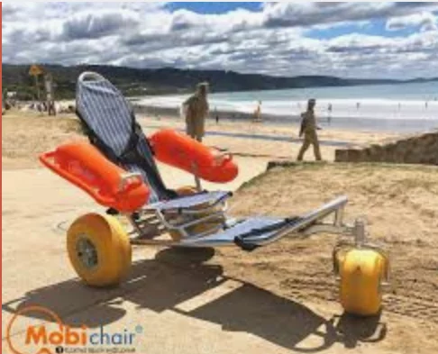 A beach wheelchair with flotation devices, designed for accessibility, parked on sandy terrain near the ocean. In the background, people are walking along the beach.