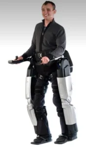 A man standing in an exoskeleton suit, smiling and holding a remote control. The suit has a sleek design and is primarily black and silver, enhancing the man's mobility.