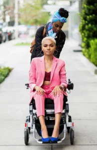 A woman in a pink suit sitting in a wheelchair, while another woman with a blue headscarf adjusts her outfit from behind. They are on a sidewalk lined with greenery in an urban area.