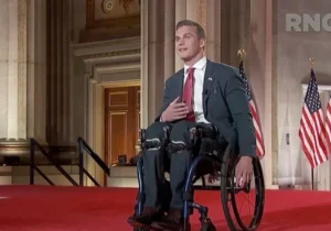 A man in a suit sitting in a wheelchair, smiling and gesturing with one hand while speaking on a red carpet stage. Behind him are American flags and a grand interior setting.