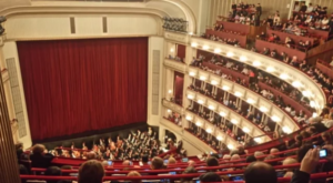 An aerial view of an opera house interior, showcasing an orchestra on stage in front of a red curtain, with a full audience in elegantly arranged seating.