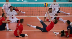 Male athletes playing sitting volleyball during a match. One player dives to hit the ball over the net while others are seated on the court, ready for the play.