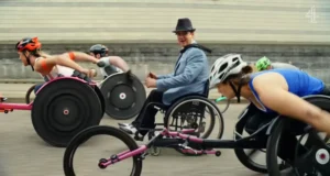 A group of athletes in racing wheelchairs competing on a track. One man in a suit and hat is smiling, while three women in bright sportswear focus on their race.