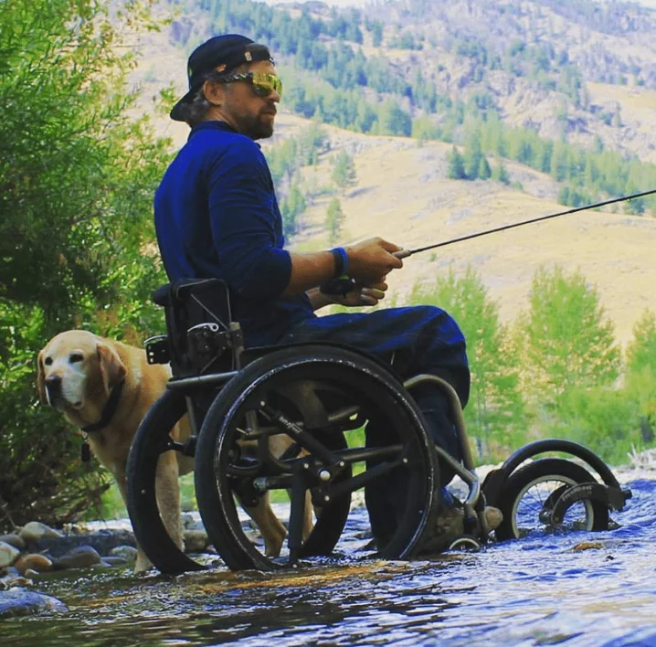 A man in a wheelchair fishing beside a river, accompanied by a yellow Labrador dog. The scene features green trees and mountains in the background, conveying a natural outdoor setting.