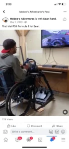 A person using a modified gaming setup with a steering wheel and seat, playing Formula 1 on a TV screen in a room. The setup includes a wheelchair adaptation for accessibility.