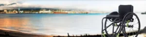 A black wheelchair positioned on a sandy beach with a scenic view of a calm sea and distant industrial area under a cloudy sky.