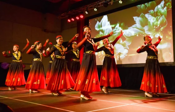 A group of dancers in red and black traditional outfits performing on stage, with a colorful floral backdrop. The scene captures the energy and vibrancy of a cultural performance, highlighting the theme of Folklorama Accessibility frequently asked questions.