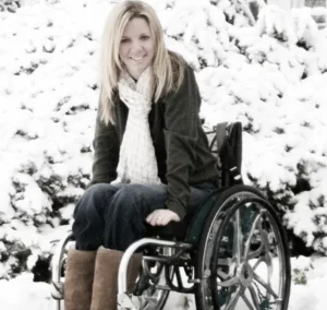 A woman with long blonde hair smiles while sitting in a wheelchair on a snowy background. She wears a green jacket, a white scarf, and brown boots, surrounded by snow-covered bushes.