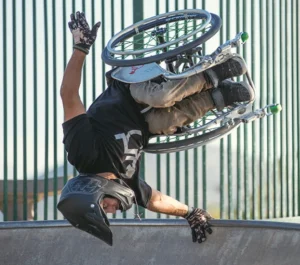 An individual performing a trick in a wheelchair at a skatepark, upside down against a green backdrop of vertical bars, wearing a helmet and gloves.
