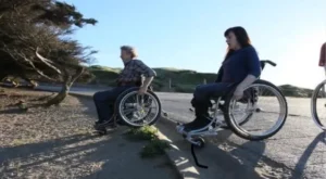 Two individuals in wheelchairs positioned near a pathway with a scenic view. One person is wearing a plaid shirt and the other is dressed in dark clothing, both facing the horizon with natural surroundings.