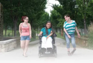 Three young women are walking on a paved path in a park. One woman in a wheelchair smiles at her friends, who are standing beside her. The surrounding area features trees and a grassy landscape.