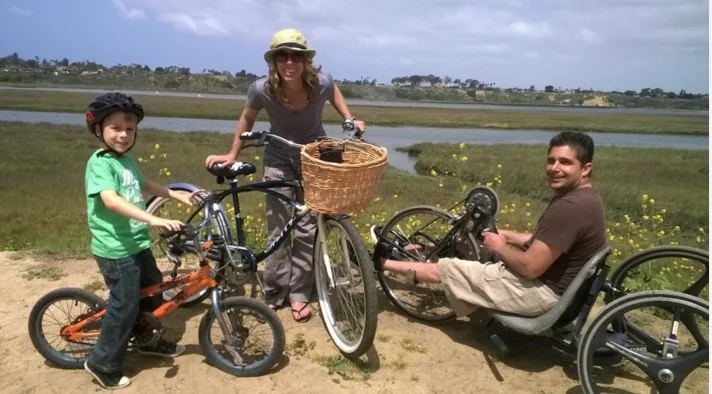 A woman standing with two bicycles, one with a basket. A young boy on an orange bike wearing a helmet stands beside her. A man in a wheelchair with hand pedals sits next to them, enjoying a scenic view of a river and grassy area.