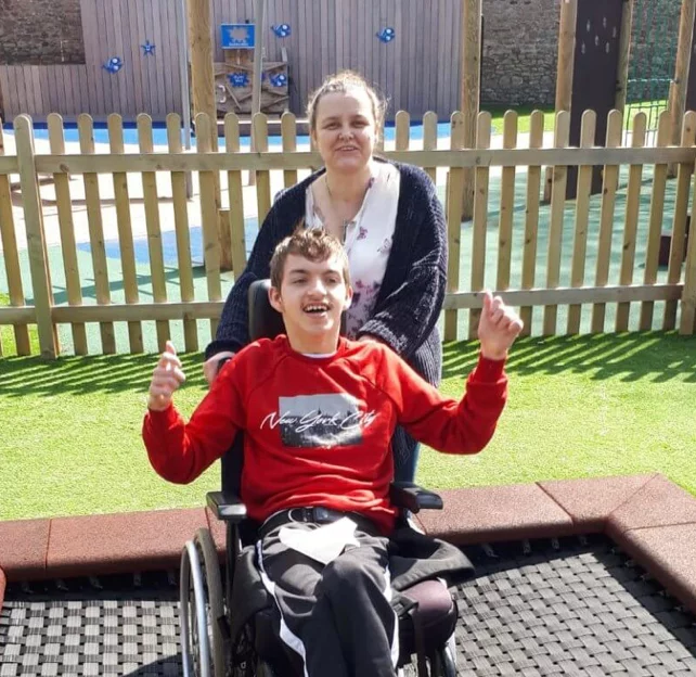 A boy wearing red shirt & sitting in wheelchair is cheering up and his mother standing by him