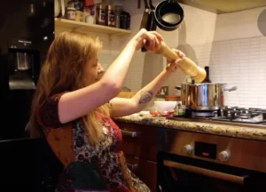 A woman is using a pepper grinder while cooking in a kitchen. She is seated at a counter with a pot on a stove, and various cooking ingredients are visible around her.