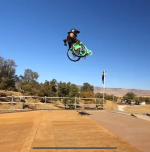 An athlete performing a trick on a wheelchair at a skatepark, soaring above a ramp with trees and a clear blue sky in the background.