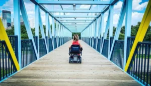 A person using a motorized wheelchair navigates a colorful pedestrian bridge in an urban park. Bright blue and yellow structural elements frame the scene, with greenery visible in the background. Accessible ways to play in Minneapolis.