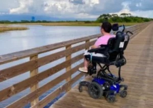 A person in a powered wheelchair is positioned on a wooden boardwalk overlooking a calm body of water. The scene features a cloudy sky and greenery in the background.