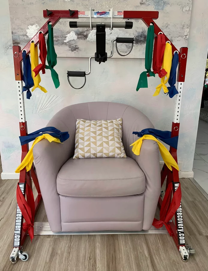 A rehabilitation exercise device placed over a light-colored armchair, featuring colorful resistance bands attached to a metallic frame. The chair has a decorative pillow on it, set against a neutral wall.
