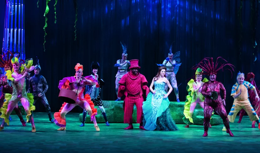 A vibrant stage performance featuring a cast in colorful costumes, showcasing aquatic-themed characters. The central performer wears a shimmering teal costume, surrounded by other actors in bright outfits, set against a dark backdrop with ocean-like lighting. Rainbow Stage Accessibility is a focus.