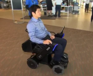 A person using a modern electric wheelchair with a sleek design, navigating through a spacious indoor area with several people in the background.