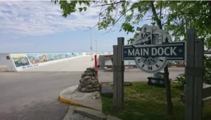 A sign reading 'MAIN DOCK' at the entrance of a waterfront area, with a view of a wide dock in the background featuring murals. Trees and a landscaped area are visible nearby.