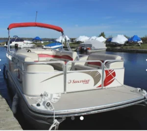 A white pontoon boat with a red canopy docked at a marina. The boat features cushioned seating and is moored next to several boats covered with blue tarps in the background.