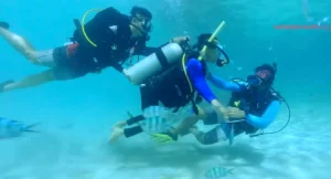 Three scuba divers underwater, one assisting another who is holding a snorkel, with various fish swimming around them. The background shows a sandy ocean floor.
