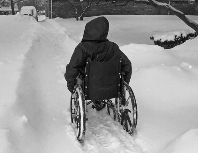 A man wearing a black hoodie, sitting in a wheelchair navigating a snowy path