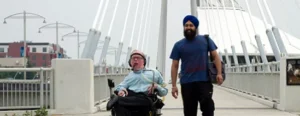 A man in a wheelchair with a helmet is accompanied by another man wearing a turban, walking on a pedestrian bridge. Both appear to be enjoying their outing in a modern urban setting.