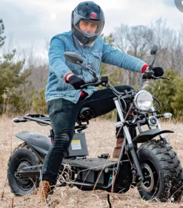 A person wearing a helmet and denim jacket sits on an electric scooter in an open field, surrounded by sparse trees, showcasing a casual outdoor ride.