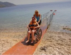 A woman assisting a person in a wheelchair on a wooden ramp leading to the water at a beach, with calm waves in the background and a clear sky.