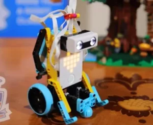 A playful robotic toy with a gray rectangular head, blue wheels, and a mix of yellow and black components, positioned on a wooden table with a blurred background of a colorful scene.