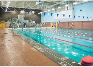 A spacious indoor swimming pool with several lanes, featuring blue pennants hanging from the ceiling. Swimmers are visible in the water, and a lifeguard chair stands poolside beside a swim lane. Wet tile and diving blocks are also present.