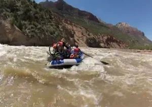 A group of people navigating a white-water raft on a river surrounded by rocky cliffs and green vegetation under a clear blue sky.