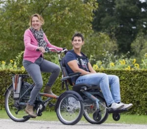 A woman riding a bicycle with a man seated in an adapted wheelchair attached to it, set against a background of greenery and sunflowers.
