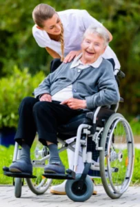 A caregiver smiling and gently supporting an elderly woman in a wheelchair, both outdoors with green foliage in the background. The elderly woman is smiling, wearing a gray cardigan.