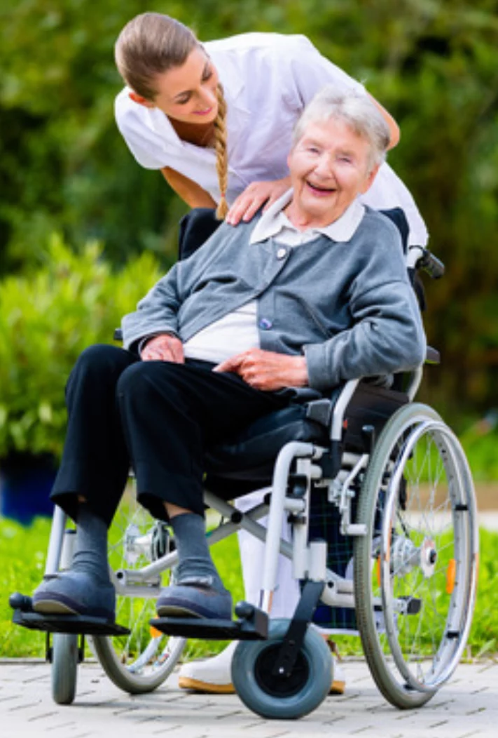 A caregiver smiling and gently supporting an elderly woman in a wheelchair, both outdoors with green foliage in the background. The elderly woman is smiling, wearing a gray cardigan.