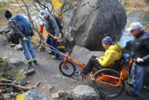 A group of four people assisting a person in a specialized wheelchair, navigating a rocky path near a river. The individual in the wheelchair is wearing a yellow jacket and a beanie, while the others are using ropes for support.
