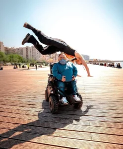 A man in a wheelchair watches as another man performs a jump nearby on a sunny walkway. The scene features a wooden boardwalk lined with people in the background and buildings in the distance.